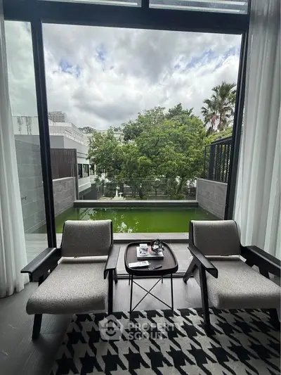 Luxurious living room with large window overlooking serene garden and pool, featuring modern chairs and stylish decor.