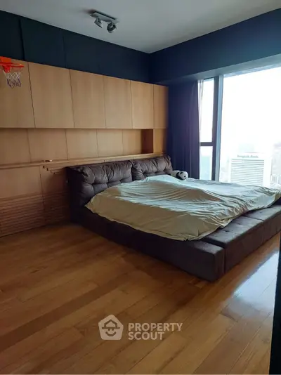 Spacious bedroom with large window offering city view and modern wooden furnishings.