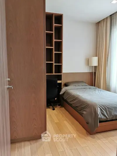 Cozy bedroom with wooden flooring, built-in shelves, and natural light from large window.
