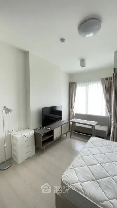 Modern bedroom with natural light, featuring a TV, cozy seating, and minimalist decor.