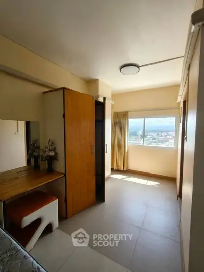 Bright bedroom with large window offering stunning city views, featuring a wooden wardrobe and a cozy dressing area.