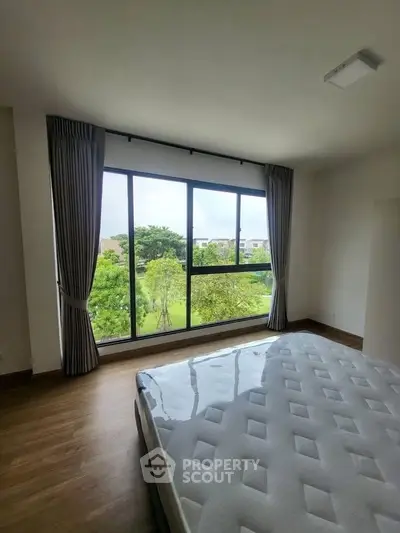 Spacious bedroom with large window offering lush green views, featuring a comfortable mattress and elegant curtains.