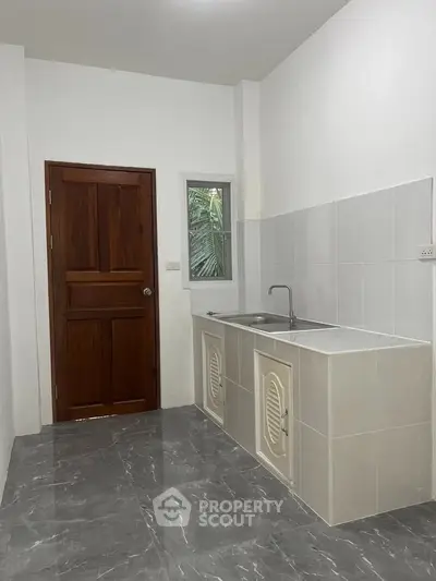 Modern kitchen with sleek tile flooring and wooden door, featuring a compact sink area and window for natural light.