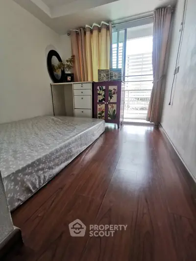 Cozy bedroom with wooden flooring and balcony access, featuring natural light and simple decor.