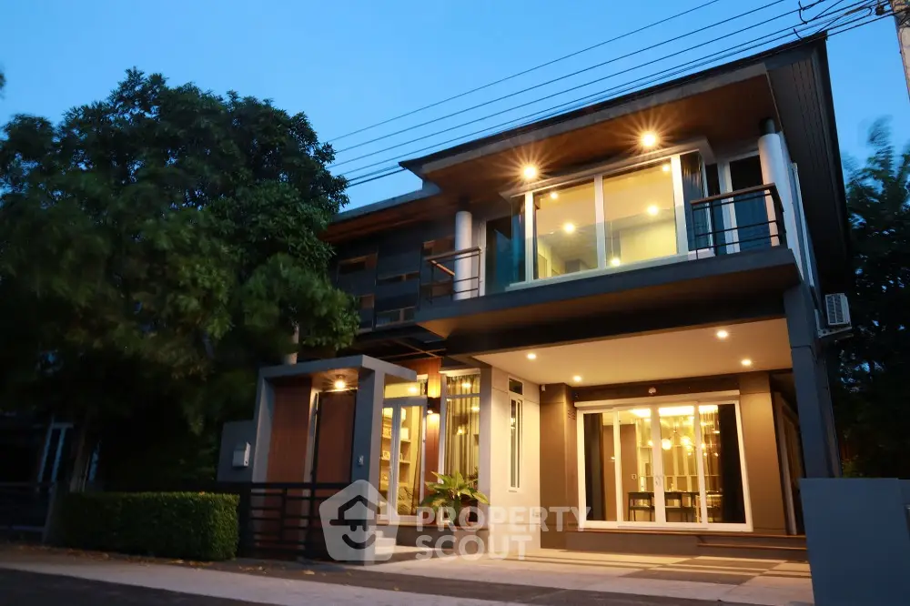 Modern two-story house with large windows and evening lighting, surrounded by lush greenery.