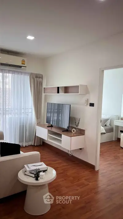 Modern living room with sleek furniture and wooden flooring, featuring a wall-mounted TV and large window.