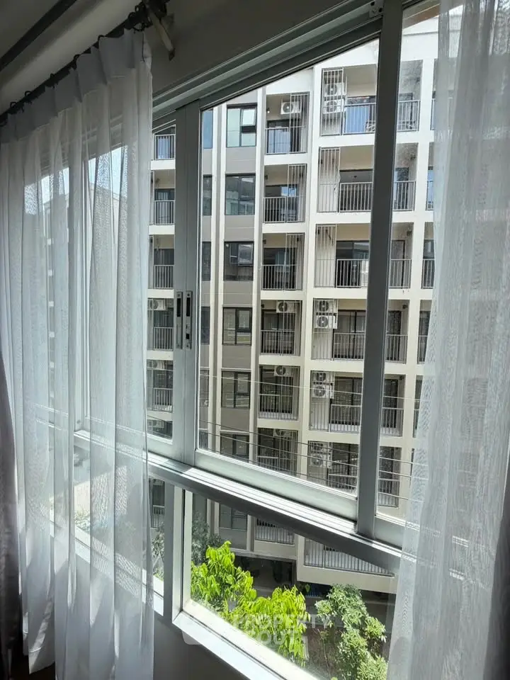 Modern apartment window view with sheer curtains overlooking neighboring building and greenery.
