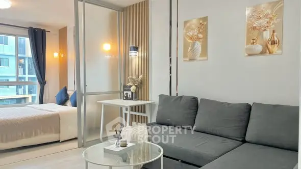 Modern living room with gray sofa and glass coffee table, adjacent to a cozy bedroom with large windows.