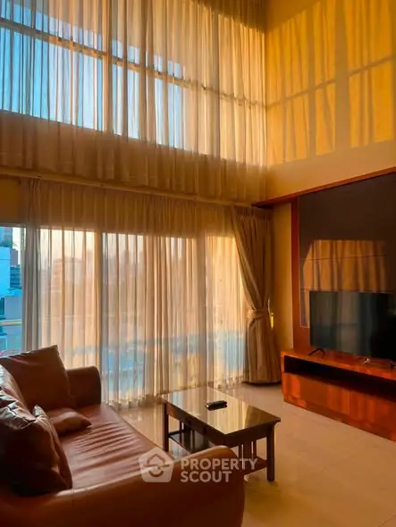 Spacious living room with floor-to-ceiling windows and elegant curtains, bathed in warm sunlight.