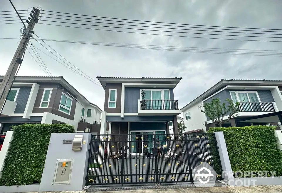 Modern two-story house with gated entrance and lush greenery under cloudy skies.