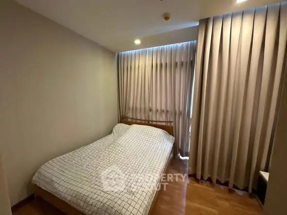 Cozy bedroom with wooden flooring and large window draped with curtains.