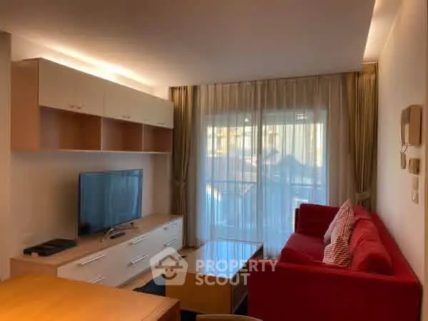 Modern living room with red sofa and large TV, featuring natural light from balcony window.