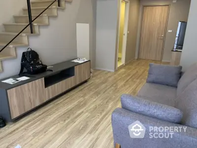 Modern living room with elegant grey sofa, sleek wooden entertainment unit, and warm wood flooring, leading to a cozy staircase and kitchen area.