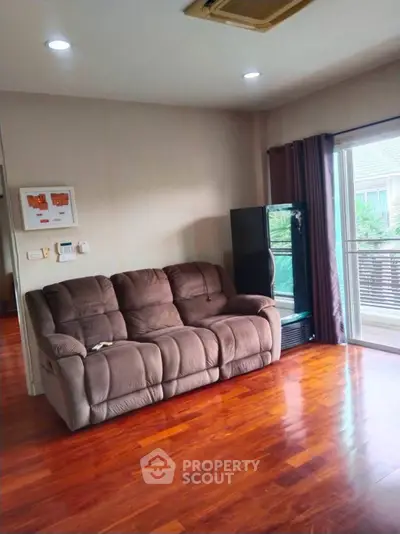 Spacious living room with plush sofa and balcony access, featuring elegant wooden flooring and ample natural light.