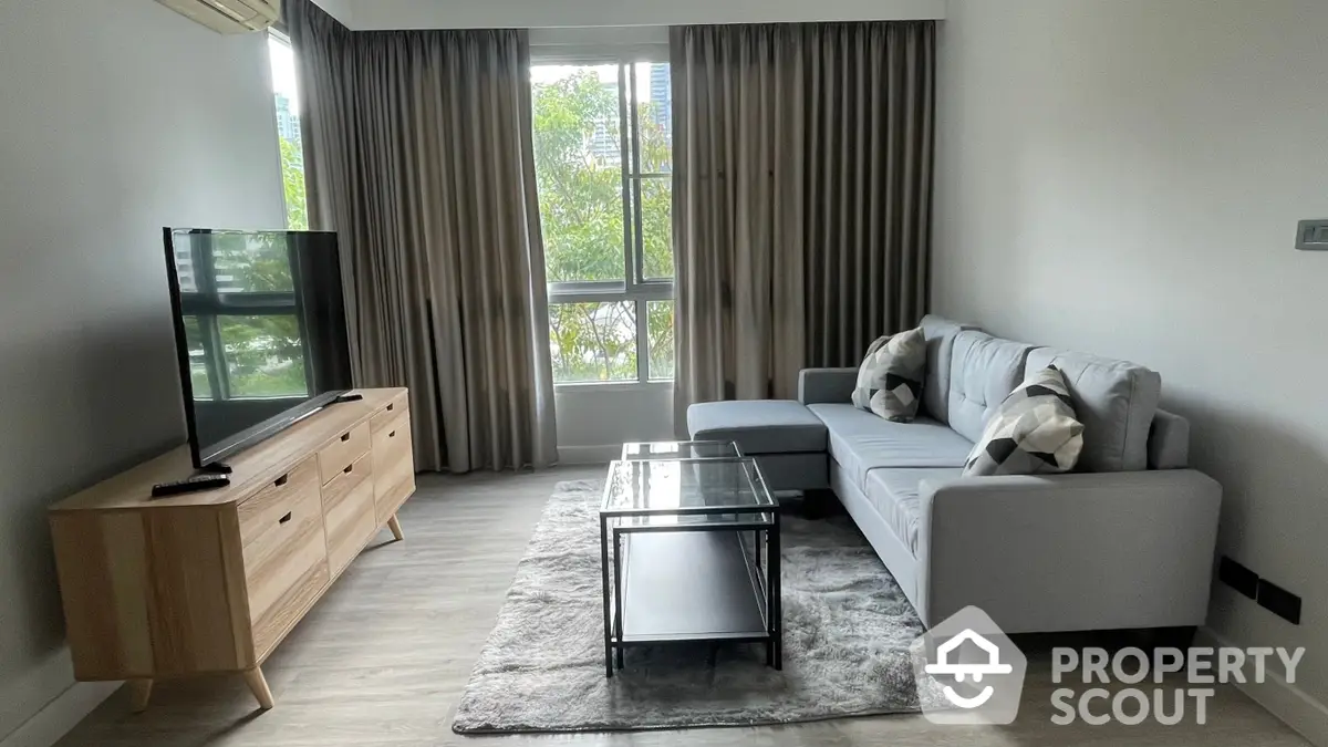 Spacious living room with modern furnishings, large windows allowing ample natural light, and a cozy grey sofa set, perfect for relaxation and entertainment.