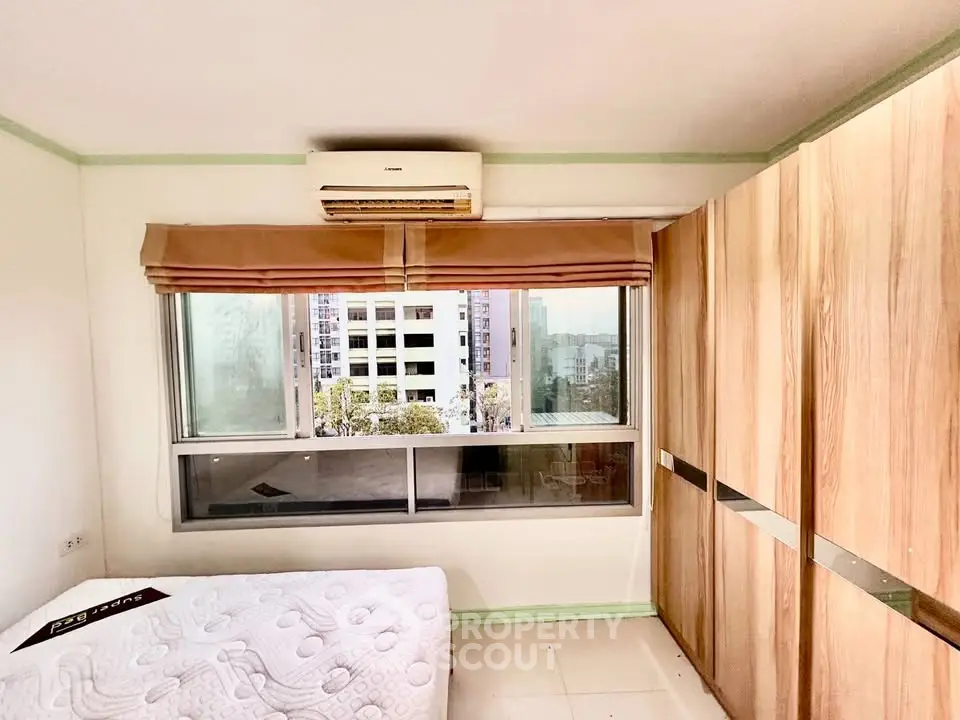 Bright bedroom with large window and city view, featuring built-in wooden wardrobe and air conditioning.