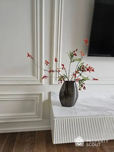 Elegant interior with classic white wall molding, a sleek flat-screen TV, and a stylish vase with vibrant flowers on a marble table, showcasing a modern yet timeless design.