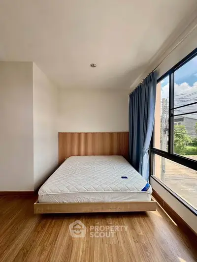 Bright bedroom with large window and wooden flooring, featuring a minimalist bed frame and blue curtains.