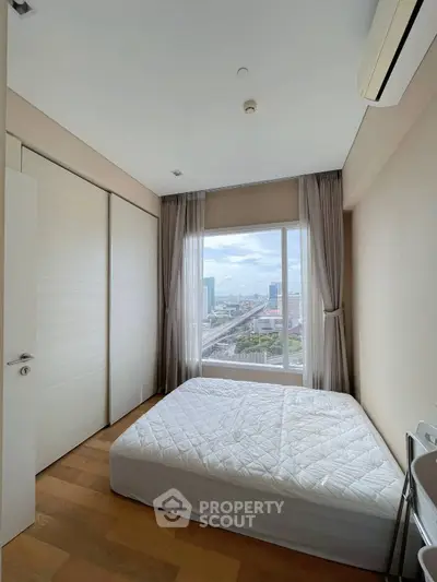 Bright bedroom with large window offering stunning city view, featuring minimalist decor and wooden flooring.