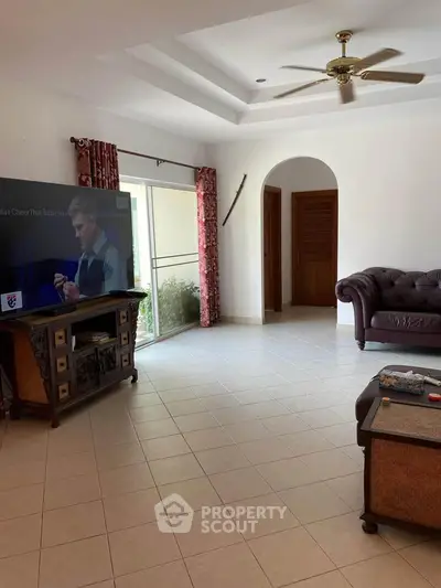 Spacious living room with elegant sofa and large TV, featuring natural light and ceiling fan.
