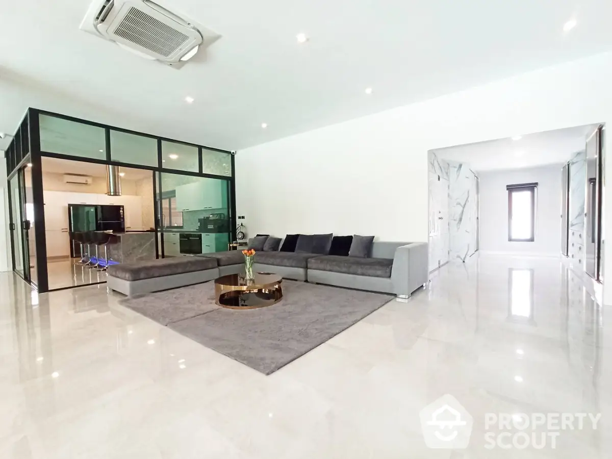 Spacious and modern living room with glossy white floors, elegant grey sectional sofa, and glass partition leading to a sleek kitchen.