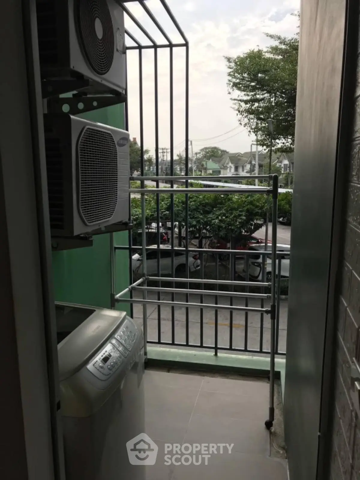 Compact balcony with washing machine and air conditioning unit, overlooking a suburban street view.