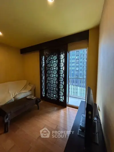 Cozy living room with wooden flooring and intricate window paneling, offering a glimpse of the balcony.