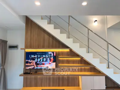 Modern living room with stylish staircase and wall-mounted TV, featuring sleek wooden paneling and ambient lighting.