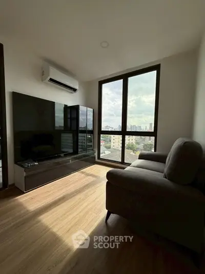 Modern living room with large windows offering city views, featuring sleek furniture and wooden flooring.