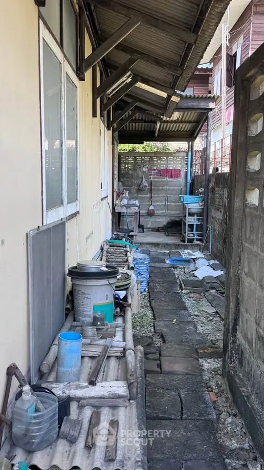 Narrow outdoor passageway with rustic charm, featuring corrugated roofing and eclectic storage solutions.