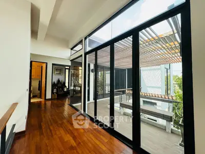 Spacious living room with polished hardwood floors extending to a sunlit balcony with modern glass barriers and a pergola, offering a seamless indoor-outdoor living experience.