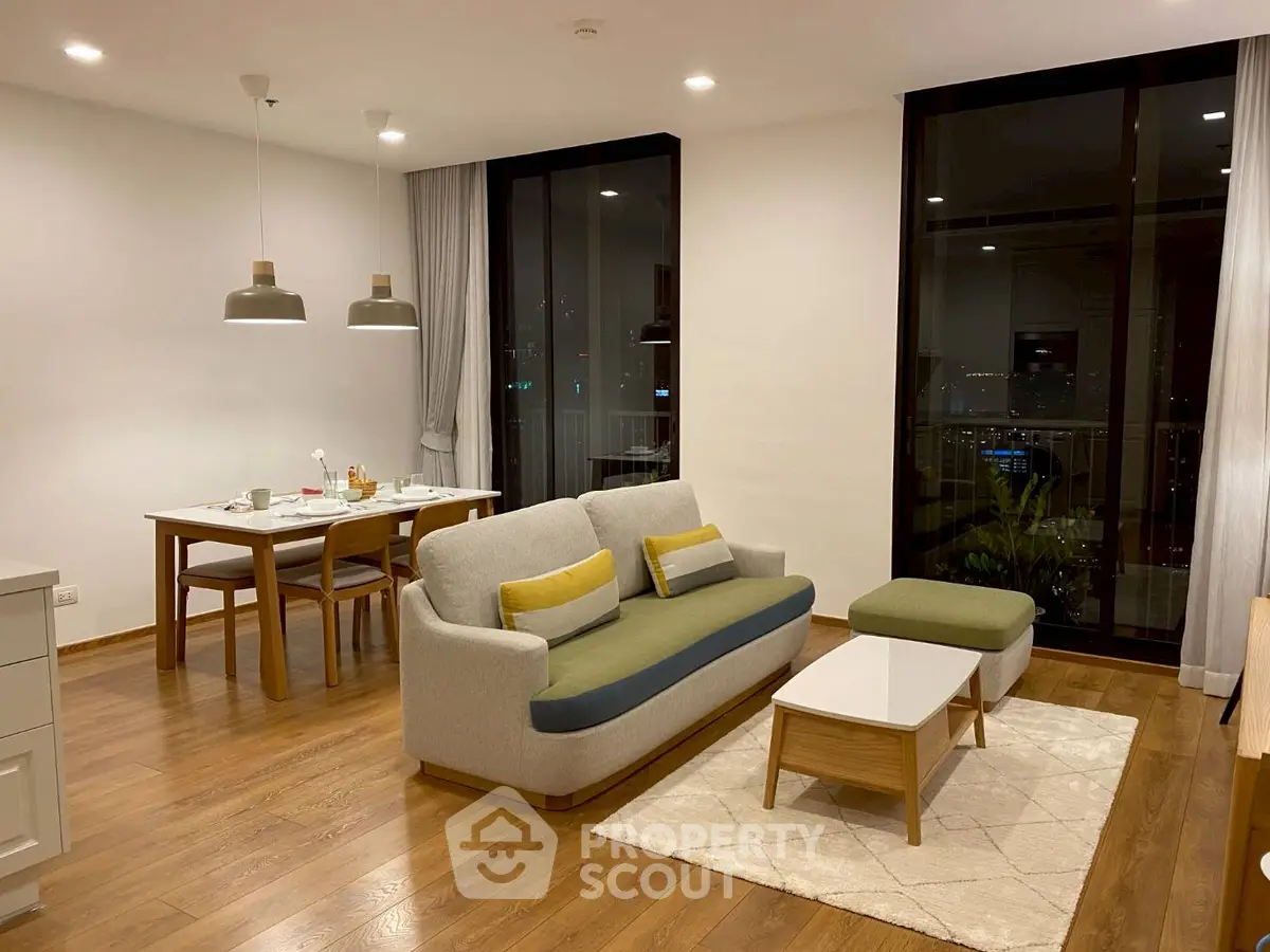 Modern living room with stylish furniture and dining area, featuring large windows and wooden flooring.