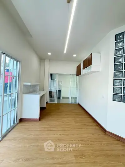 Spacious modern living room with wooden flooring and glass doors leading to a bright kitchen area.