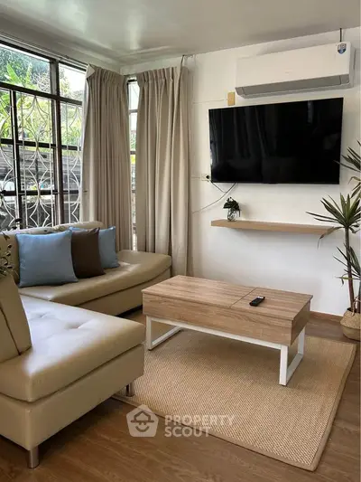 Modern living room with sleek furniture, wall-mounted TV, and large windows offering natural light.