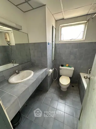 Compact bathroom with gray tiles, featuring a sink, toilet, and bathtub under natural light.