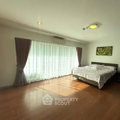 Spacious bedroom with large windows and elegant curtains, featuring a cozy bed and wooden flooring.