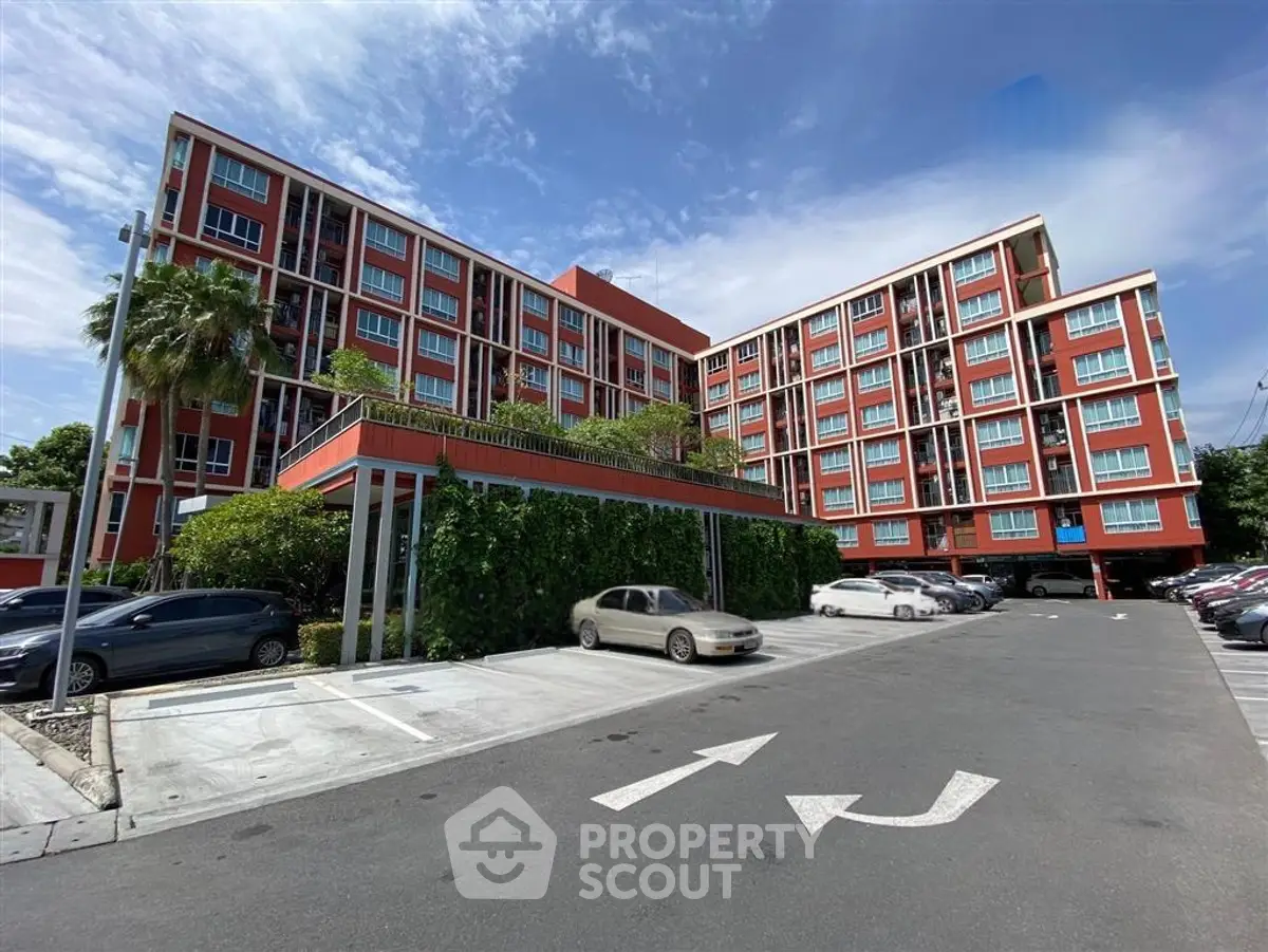 Modern red apartment building with lush greenery and spacious parking area under a clear blue sky.