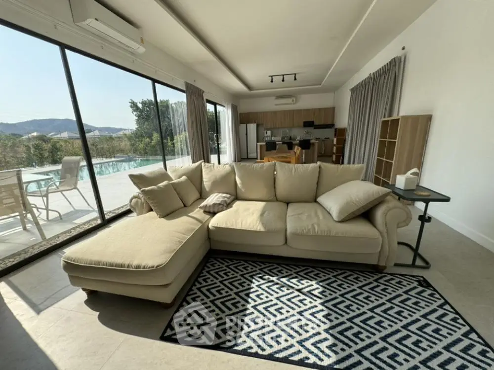 Spacious living room with large sectional sofa and geometric rug, opening to a sunlit balcony with a serene pool view, perfect for luxurious relaxation.
