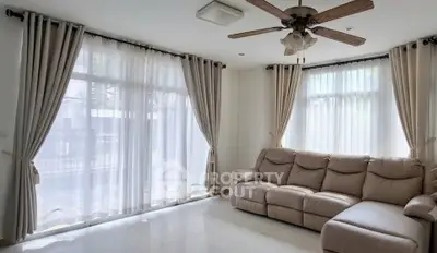 Spacious living room with large windows and elegant curtains, featuring a comfortable beige sofa and ceiling fan.