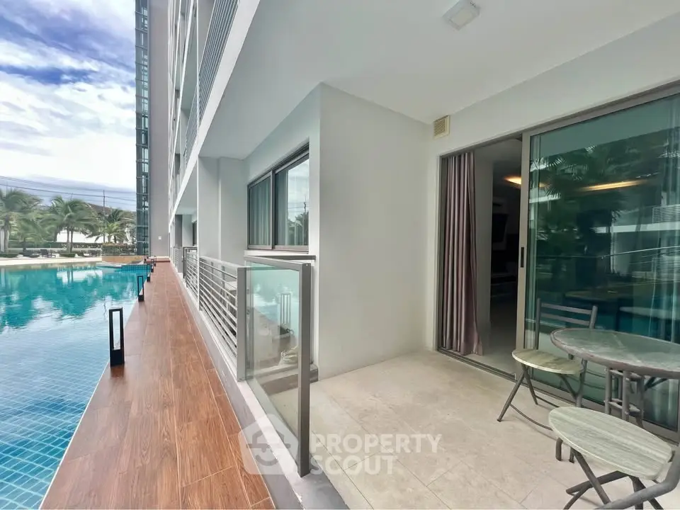Modern poolside balcony with glass railing and outdoor seating in luxury apartment complex.