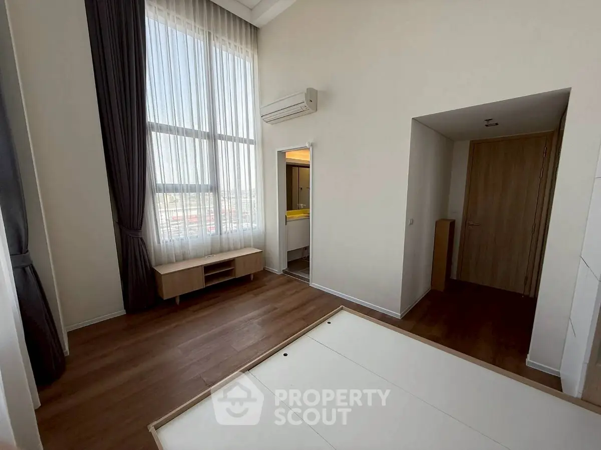 Spacious bedroom with high ceilings and large window, featuring modern wooden flooring and air conditioning.