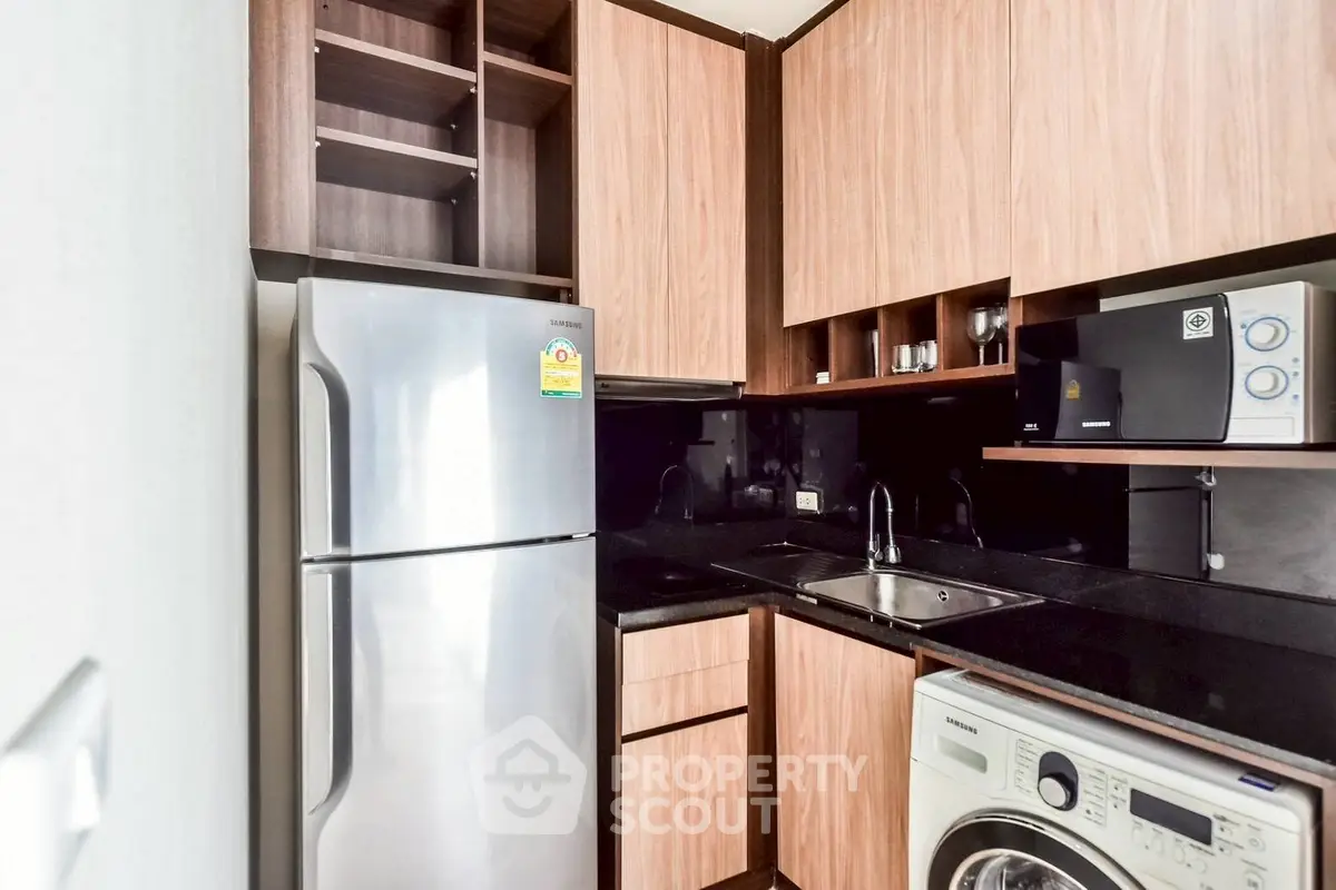 Modern kitchen with stainless steel fridge and washing machine, sleek wooden cabinets, and black countertops.
