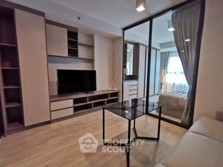 Modern living room with glass table and TV, adjacent to cozy bedroom with natural light.