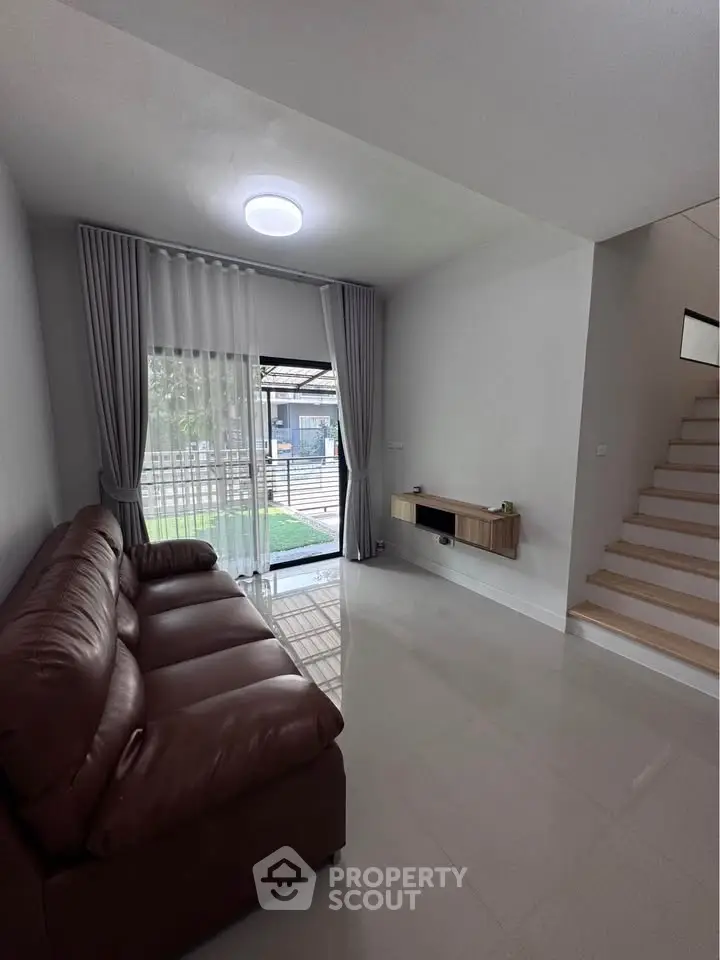 Modern living room with leather sofa and garden view, featuring sleek staircase and natural light.