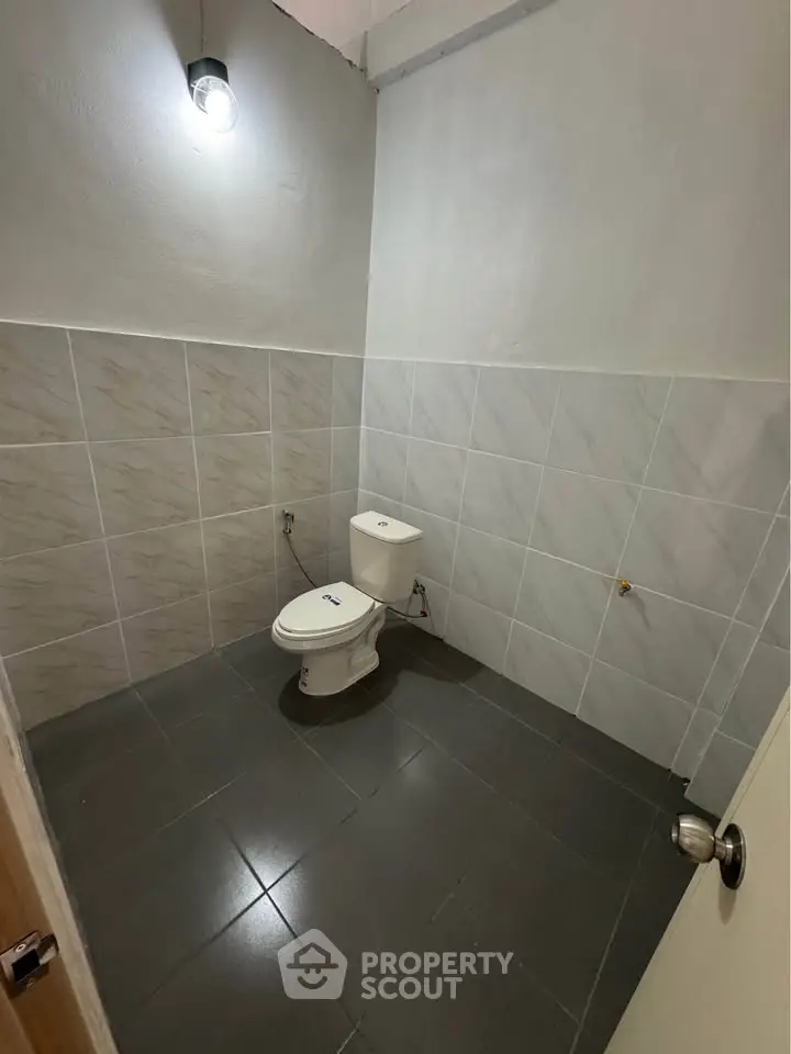 Minimalist bathroom with tiled walls and floor, featuring a single toilet under a wall-mounted light.
