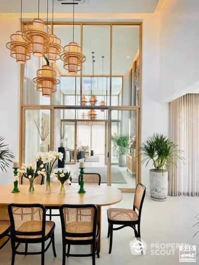 Elegant dining space with rattan chairs, chic pendant lights, and a view into a serene sunroom, surrounded by lush greenery and natural light.