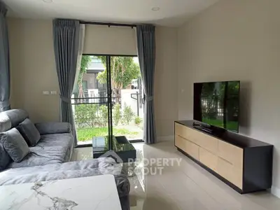 Modern living room with large sofa, flat-screen TV, and garden view through sliding glass doors.
