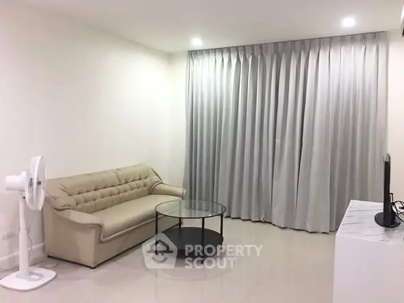 Modern living room with beige sofa, glass coffee table, and floor fan in a minimalist setting.