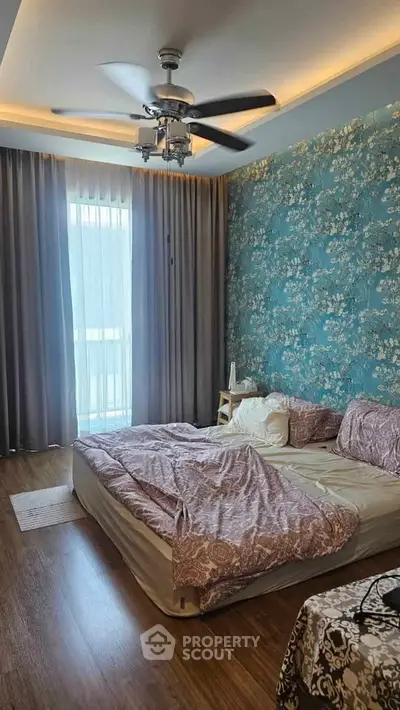Charming bedroom with floral accent wall and ceiling fan, featuring cozy bedding and natural light from large window.