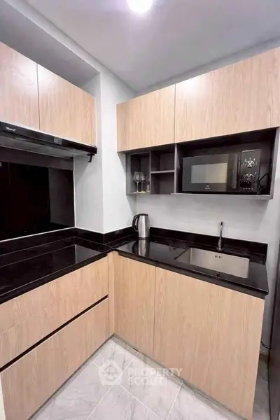 Modern kitchen with sleek black countertops and light wood cabinets, featuring a microwave and electric kettle.